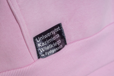 Naszywka z nazwą Uniwersytetu Kazimierza Wielkiego w Bydgoszczy na różowej bluzie.