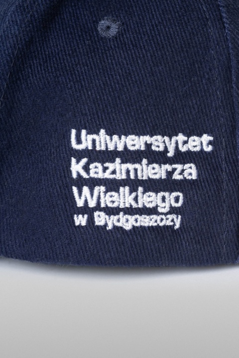 Zbliżenie białego haftu z nazwą Uniwersytetu na granatowej czapce.