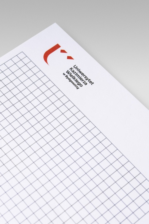 Zbliżenie górnej części strony notatnika w kratkę z czerwonym znakiem graficznym i czarnym napisem „Uniwersytet Kazimierza Wielkiego w Bydgoszczy”.
