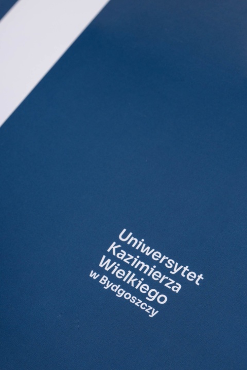 Zbliżenie niebieskiej okładki teczki z białym napisem „Uniwersytet Kazimierza Wielkiego w Bydgoszczy” i fragmentem białego znaku graficznego.