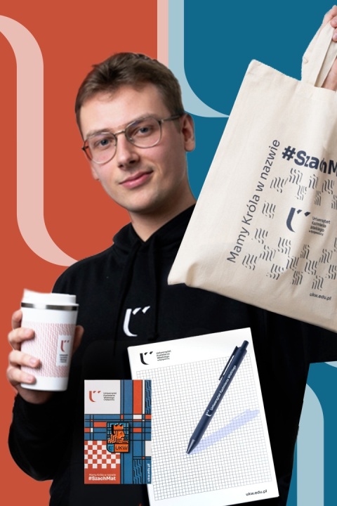 Mężczyzna w czarnej bluzie UKW prezentuje Zestaw na start premium: kubek termiczny, torbę, notes A4, długopis i pin.
